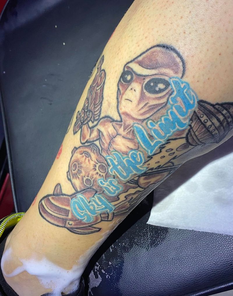 Color tattoo of a cartoon alien with a blue script banner on a forearm.