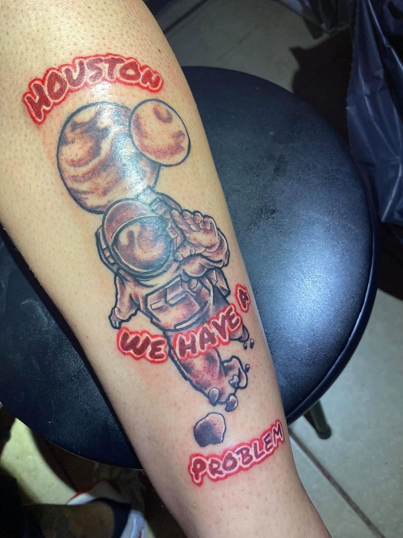 Color tattoo of a cartoon astronaut holding balloons with the red lettering Houston We Have a Problem on a forearm.