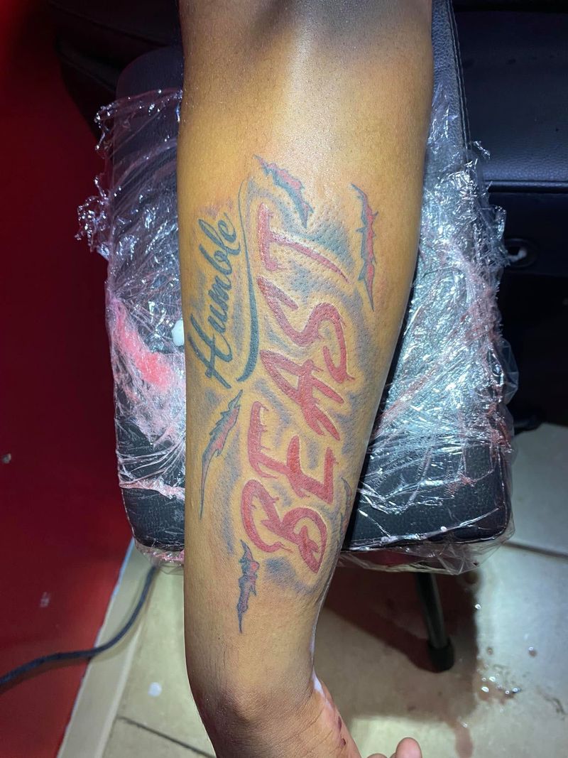 Lettering tattoo reading Humble Beast in green script and bold red letters running down a forearm.