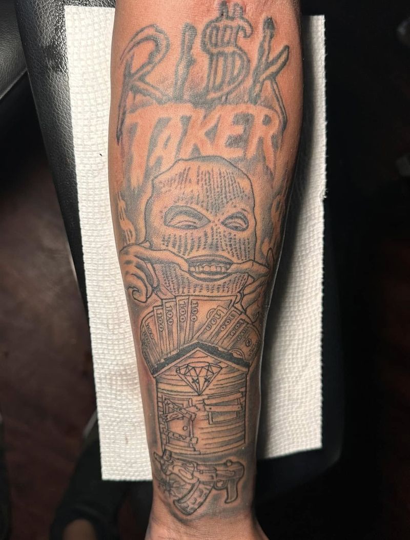 Black and grey forearm tattoo with the words Risk Taker above a masked figure and stacked imagery below.